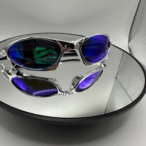 Op de afbeelding: Zilverkleurige zonnebril met blauwe glazen, gepresenteerd op een reflecterend oppervlak. De zonnebril heeft een modern design met een metallic afwerking. De glazen reflecteren een groene tint, wat een levendig contrast cre&euml;ert. De afbeelding is goed belicht en toont de details van de bril.