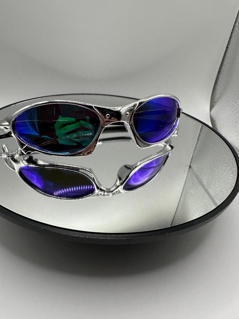 Op de afbeelding: Zilverkleurige zonnebril met blauwe glazen, gepresenteerd op een reflecterend oppervlak. De zonnebril heeft een modern design met een metallic afwerking. De glazen reflecteren een groene tint, wat een levendig contrast cre&euml;ert. De afbeelding is goed belicht en toont de details van de bril.