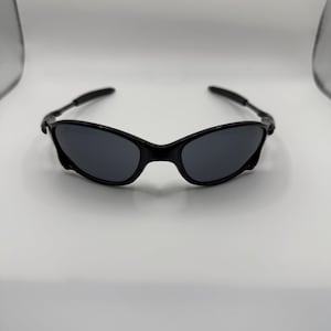 May include: Black sunglasses with dark gray lenses. The sunglasses have a sleek, curved frame design and are set against a white background. The temples are thin and extend to the ear pieces.