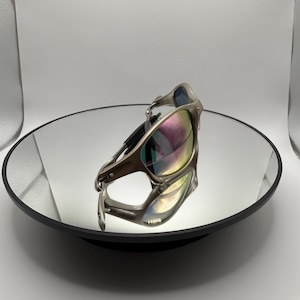 May include: Silver-framed sunglasses with iridescent lenses are displayed on a mirrored surface. The sunglasses have a sleek, modern design with a curved frame and dark temples. The lenses reflect a spectrum of colors.