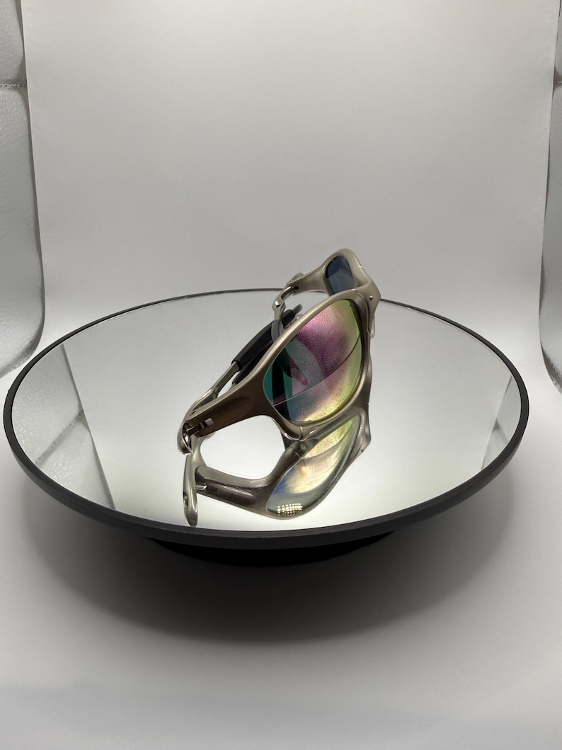 May include: Silver-framed sunglasses with iridescent lenses are displayed on a mirrored surface. The sunglasses have a sleek, modern design with a curved frame and dark temples. The lenses reflect a spectrum of colors.