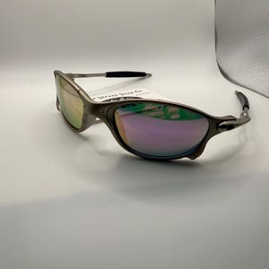 May include: A pair of silver-framed sunglasses with iridescent lenses. The sunglasses have a sleek, modern design with a curved frame and black accents on the temples. A tag is attached to the frame.