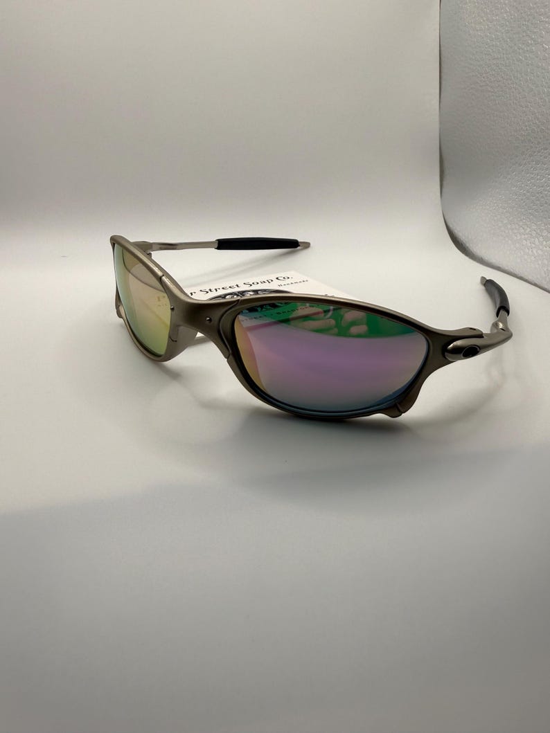 May include: A pair of silver-framed sunglasses with iridescent lenses. The sunglasses have a sleek, modern design with a curved frame and black accents on the temples. A tag is attached to the frame.