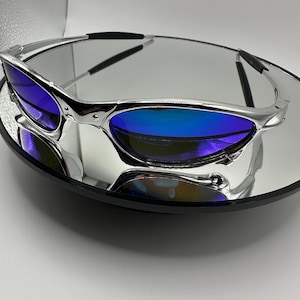 Op de afbeelding: Zilverkleurige zonnebril met blauwe glazen. De bril wordt gepresenteerd op een reflecterend oppervlak, wat het strakke ontwerp benadrukt. De glazen hebben een kleureffect, van diepblauw naar een lichtere tint.