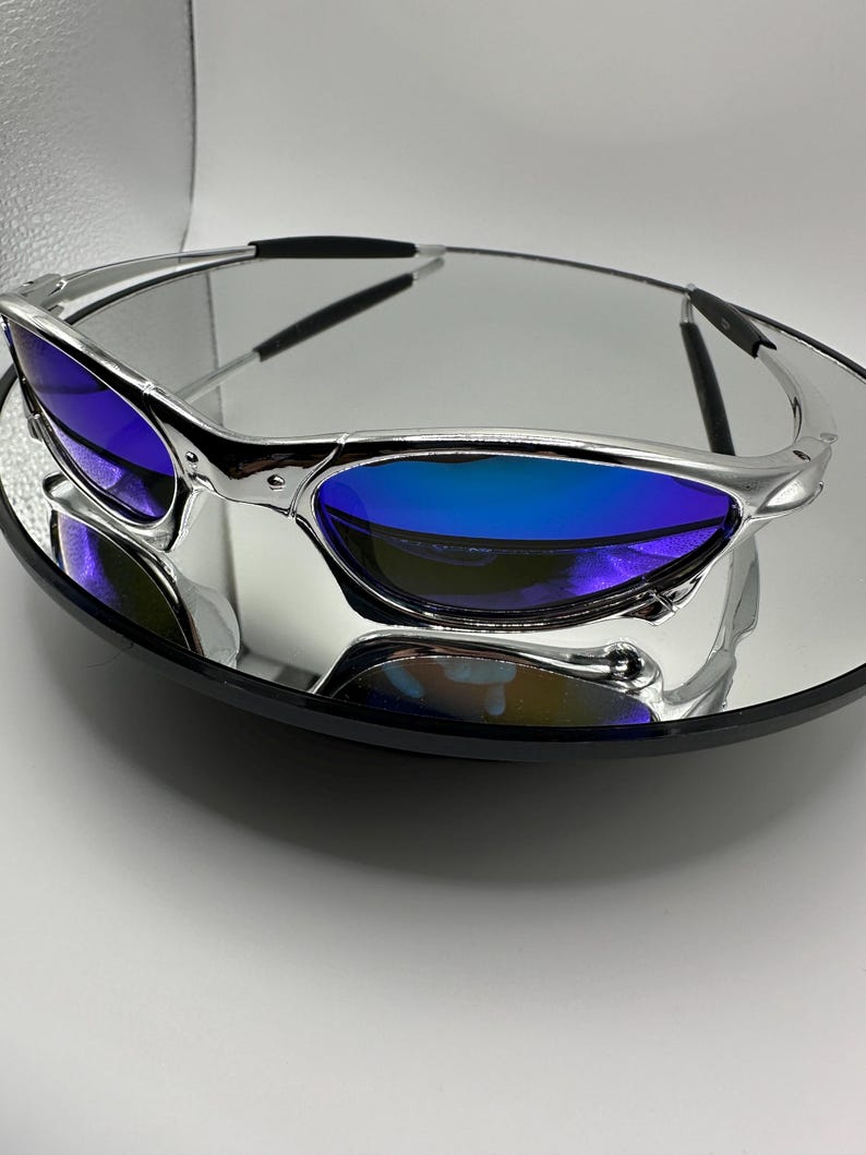 Op de afbeelding: Zilverkleurige zonnebril met blauwe glazen. De bril wordt gepresenteerd op een reflecterend oppervlak, wat het strakke ontwerp benadrukt. De glazen hebben een kleureffect, van diepblauw naar een lichtere tint.