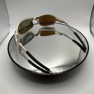 May include: A pair of silver-framed sunglasses with orange-tinted lenses, resting on a reflective surface. The sunglasses have a sleek, modern design with black temple tips. The image is well-lit, highlighting the metallic finish.