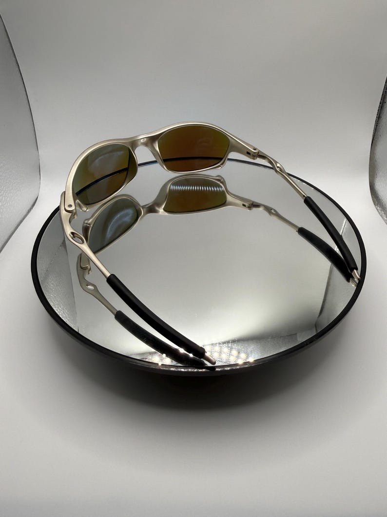 May include: A pair of silver-framed sunglasses with orange-tinted lenses, resting on a reflective surface. The sunglasses have a sleek, modern design with black temple tips. The image is well-lit, highlighting the metallic finish.
