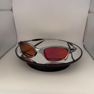 May include: Silver-framed sunglasses with red-tinted lenses. The sunglasses are displayed on a reflective black surface, set against a white background. The lenses have a gradient effect, transitioning from orange to red.