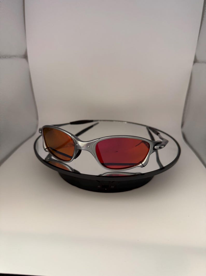May include: Silver-framed sunglasses with red-tinted lenses. The sunglasses are displayed on a reflective black surface, set against a white background. The lenses have a gradient effect, transitioning from orange to red.