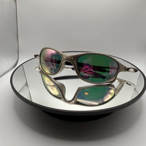 May include: Silver-framed sunglasses with green-tinted lenses are displayed on a reflective surface. The lenses reflect a rainbow of colors. The sunglasses have a sleek, modern design.