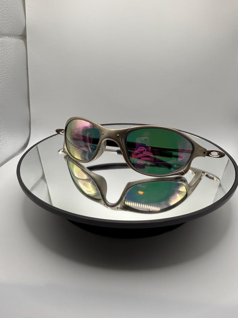May include: Silver-framed sunglasses with green-tinted lenses are displayed on a reflective surface. The lenses reflect a rainbow of colors. The sunglasses have a sleek, modern design.