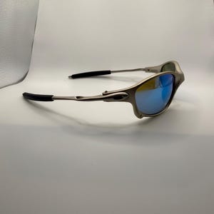 May include: Silver-framed sunglasses with blue mirrored lenses. The sunglasses have black temple tips and a sleek, modern design. The lenses reflect a vibrant blue color.