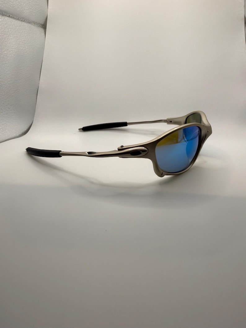 May include: Silver-framed sunglasses with blue mirrored lenses. The sunglasses have black temple tips and a sleek, modern design. The lenses reflect a vibrant blue color.