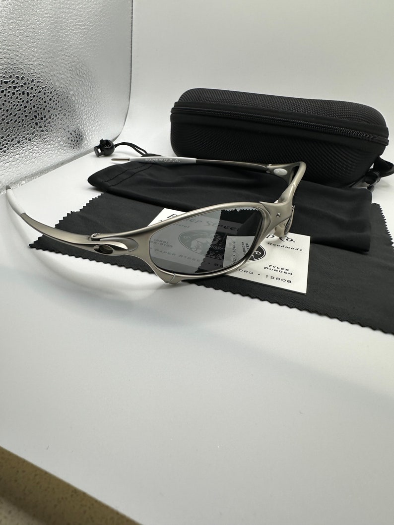 May include: Silver-toned sunglasses with a black case, black microfiber cloth, and a white tag. The sunglasses have a curved design and dark lenses. The case is rectangular and has a zipper. The tag has the text "PAPER STREET" and "TYLER DURDEN".
