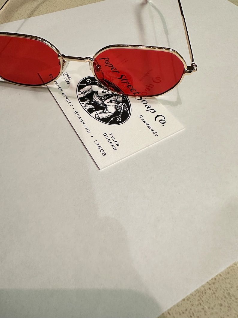 Może przedstawiać: Okulary przeciwsłoneczne w kształcie owalu, w kolorze czerwonym, z pozłacaną metalową oprawką. Okulary leżą na wizyt&oacute;wce z napisem "Paper Street Soap Co. Handmade" i danymi kontaktowymi Tylera Durdena.