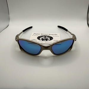 May include: A pair of metallic-framed sunglasses with blue mirrored lenses. The glasses have black earpieces and are resting on a white surface. A small card with the text "Paper Street Soap Co." is behind the glasses.