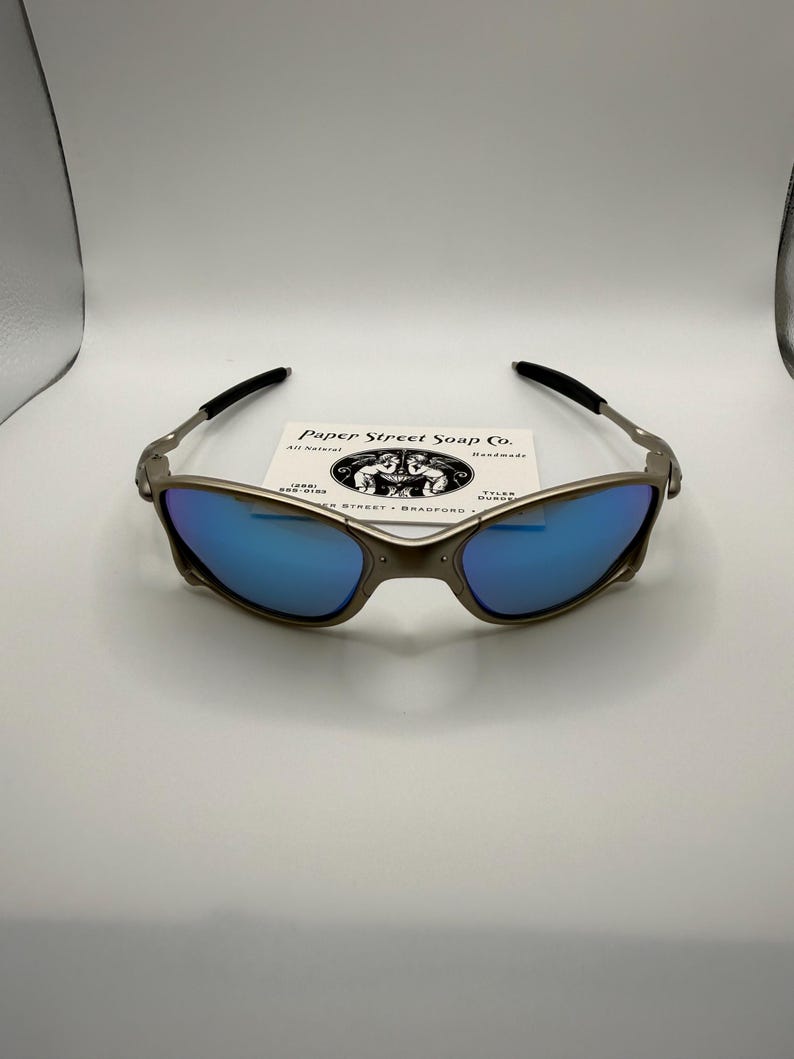 May include: A pair of metallic-framed sunglasses with blue mirrored lenses. The glasses have black earpieces and are resting on a white surface. A small card with the text "Paper Street Soap Co." is behind the glasses.