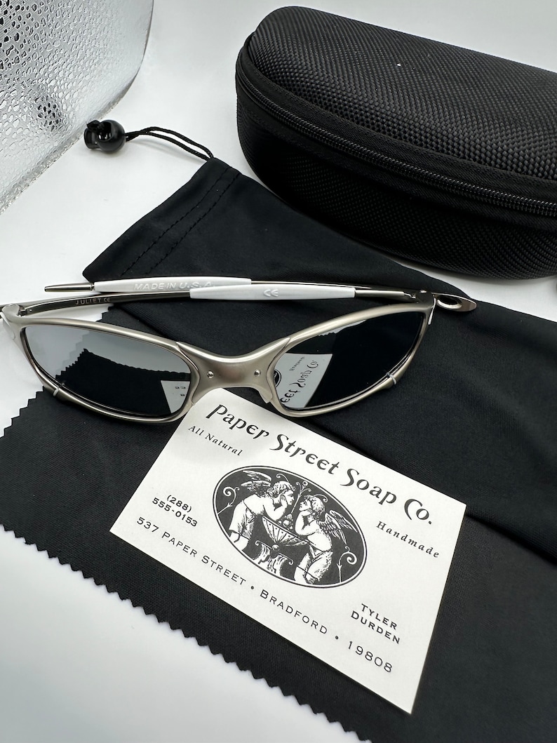 May include: Silver-framed sunglasses with dark lenses, a black case, and a black microfiber cloth. The sunglasses have a white accent on the frame. A business card for Paper Street Soap Co. is also present.