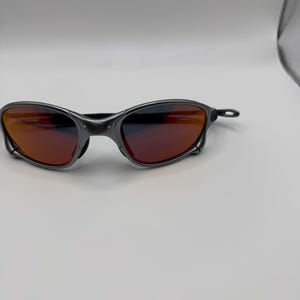 May include: A pair of sunglasses with a dark gray frame and red-tinted lenses. The sunglasses have a sleek, modern design, with a curved frame and a minimalist aesthetic. The lenses appear to be reflective.