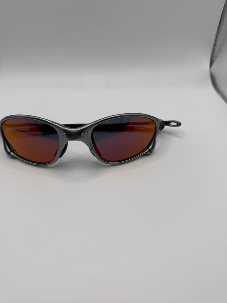 May include: A pair of sunglasses with a dark gray frame and red-tinted lenses. The sunglasses have a sleek, modern design, with a curved frame and a minimalist aesthetic. The lenses appear to be reflective.