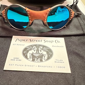 May include: A pair of blue mirrored sunglasses with a brown leather frame and stitching. The sunglasses are sitting on a black fabric surface with a business card that reads "Paper Street Soap Co. All Natural Handmade (288) 555-0153 TYLER DURDEN 537 PAPER STREET BRADFORD 19808".