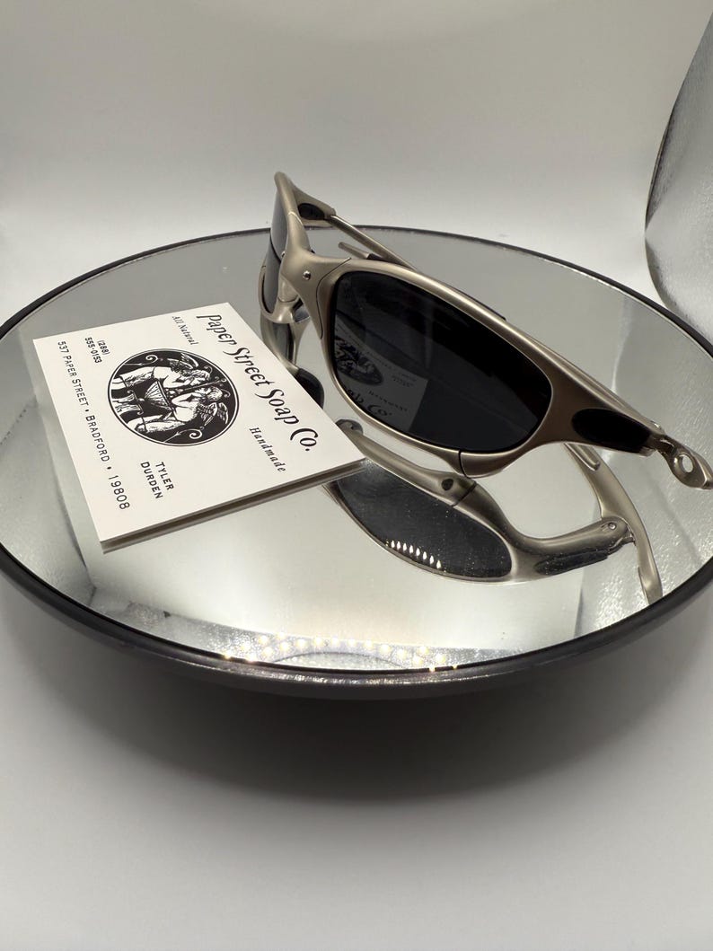 Peut inclure: Des lunettes de soleil &agrave; monture argent&eacute;e et verres fonc&eacute;s reposent sur une surface miroir. Une carte de visite pour "Paper Street Soap Co." est &eacute;galement sur le miroir. Les lunettes de soleil ont un design &eacute;l&eacute;gant et moderne.