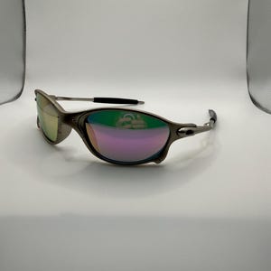 May include: A pair of metallic gray sunglasses with iridescent lenses reflecting green and purple hues. The sunglasses have a sleek, modern design with a curved frame and black accents on the temples. The lenses are oval-shaped.
