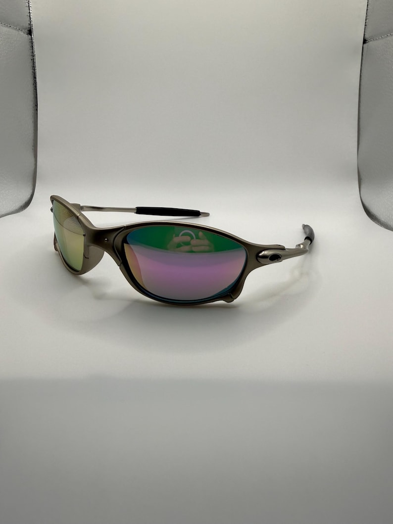 May include: A pair of metallic gray sunglasses with iridescent lenses reflecting green and purple hues. The sunglasses have a sleek, modern design with a curved frame and black accents on the temples. The lenses are oval-shaped.