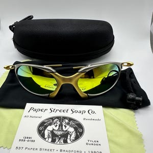 Puede incluir: Gafas de sol doradas con lentes verdes, una funda de transporte negra, una bolsa de cordón negro y una tarjeta de visita blanca con el texto "Paper Street Soap Co. All Natural Handmade (288) 555-0153 TYLER DURDEN 537 PAPER STREET BRADFORD 19808"