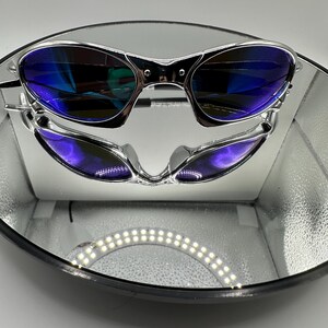 Op de afbeelding: Zilverkleurige zonnebril met blauw getinte glazen wordt gepresenteerd op een reflecterend oppervlak. De zonnebril heeft een strak, modern design. De glazen reflecteren een levendige blauwe tint en het montuur heeft een gepolijste, metalen afwerking.