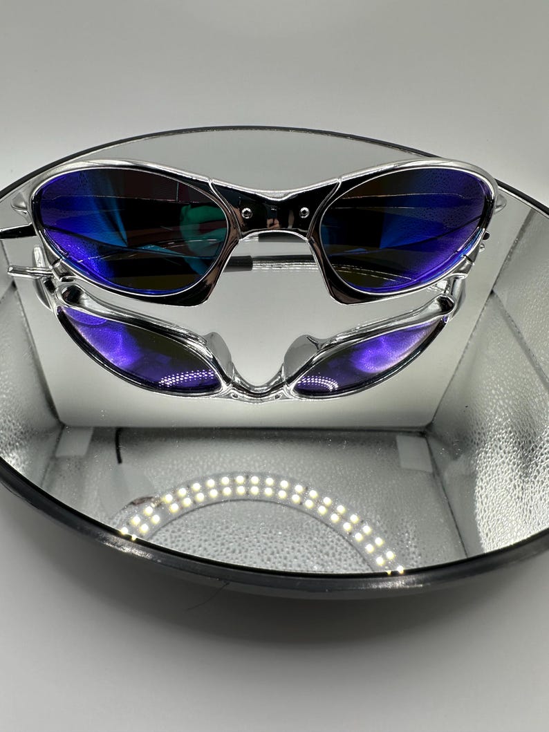 Op de afbeelding: Zilverkleurige zonnebril met blauw getinte glazen wordt gepresenteerd op een reflecterend oppervlak. De zonnebril heeft een strak, modern design. De glazen reflecteren een levendige blauwe tint en het montuur heeft een gepolijste, metalen afwerking.