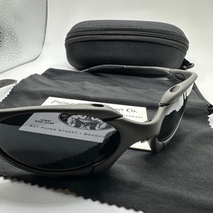 May include: A pair of black Oakley sunglasses with a gray lens. The sunglasses are sitting on a black microfiber cleaning cloth. The sunglasses are in a black case with a zipper.