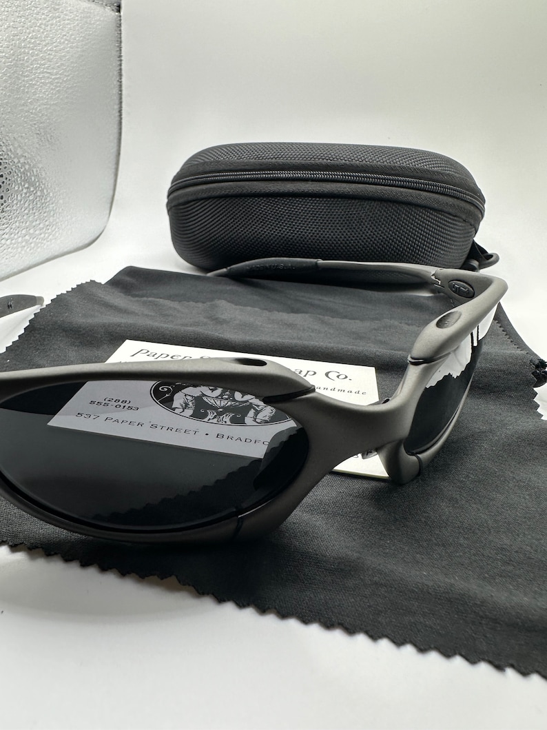 May include: A pair of black Oakley sunglasses with a gray lens. The sunglasses are sitting on a black microfiber cleaning cloth. The sunglasses are in a black case with a zipper.
