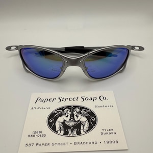 May include: Silver-framed sunglasses with blue mirrored lenses. The glasses are set against a white background, with a business card from Paper Street Soap Co. below. The card features a black and white illustration and text.
