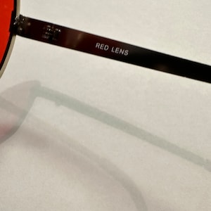 Puede incluir: Primer plano de unas gafas de sol con lentes rojos. Las gafas tienen una montura de metal negro y las palabras "RED LENS" est&aacute;n grabadas en la varilla.