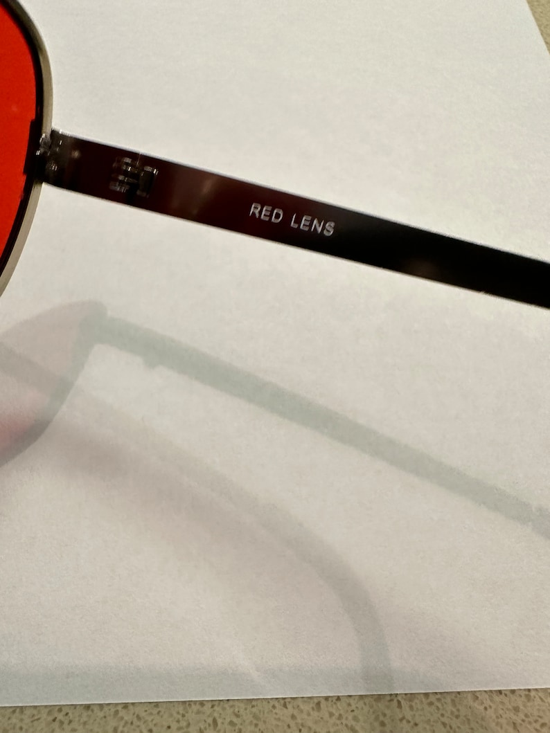 Puede incluir: Primer plano de unas gafas de sol con lentes rojos. Las gafas tienen una montura de metal negro y las palabras "RED LENS" est&aacute;n grabadas en la varilla.