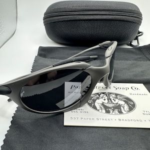 May include: A pair of gray sunglasses with a black case and a cleaning cloth. The sunglasses have a sporty design and are made of metal. The case is made of a soft, black material. The cleaning cloth is made of a soft, gray material.