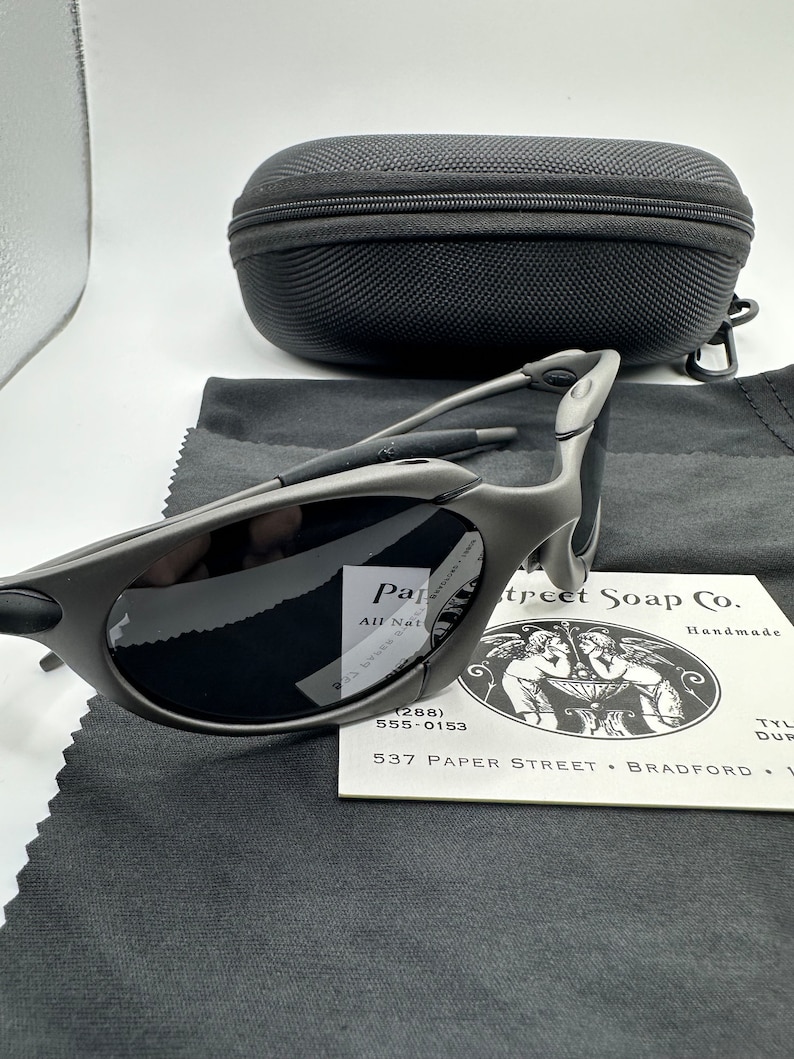 May include: A pair of gray sunglasses with a black case and a cleaning cloth. The sunglasses have a sporty design and are made of metal. The case is made of a soft, black material. The cleaning cloth is made of a soft, gray material.