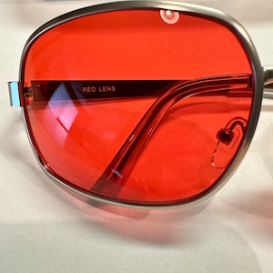 Puede incluir: Gafas de sol con montura plateada y lentes rojos. Las gafas tienen una forma redondeada y las lentes est&aacute;n tintadas de rojo. El texto "RED LENS" est&aacute; impreso en la montura.
