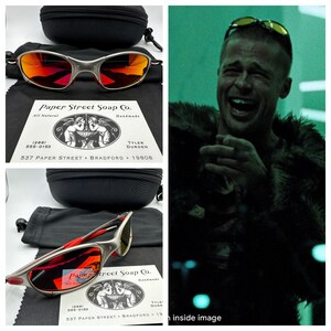 K&ouml;nnte beinhalten: Eine Sonnenbrille mit roten Spiegelgl&auml;sern, silbernem Gestell und roten Akzenten. Die Sonnenbrille befindet sich in einem schwarzen Etui mit einem schwarzen Beutel. Das Etui tr&auml;gt ein Etikett mit der Aufschrift "Paper Street Soap Co. All Natural Handmade Tyler Durden 537 Paper Street Bradford 19808".