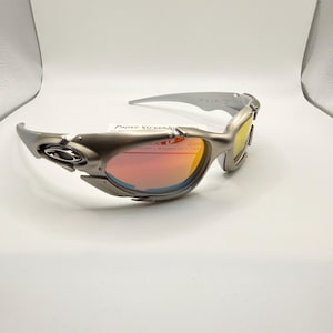 May include: Silver-framed sunglasses with a wraparound design. The lenses have a gradient of colors, transitioning from red to yellow. The text "Paper Street" is visible on the lens. The sunglasses have a sporty, futuristic aesthetic.