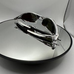 Op de afbeelding: Zonnebril met zilveren montuur en donkere glazen op een reflecterend oppervlak. De zonnebril heeft een strak, modern design met een gebogen montuur. De afbeelding is goed verlicht.