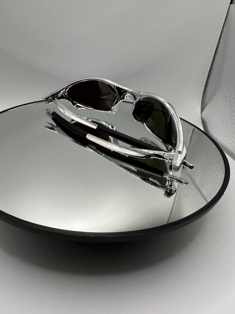 Op de afbeelding: Zonnebril met zilveren montuur en donkere glazen op een reflecterend oppervlak. De zonnebril heeft een strak, modern design met een gebogen montuur. De afbeelding is goed verlicht.