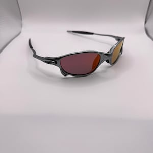 May include: A pair of sunglasses with a dark gray frame and tinted lenses. The lenses transition from a reddish-purple to a yellow-orange hue. The sunglasses are set against a white background.