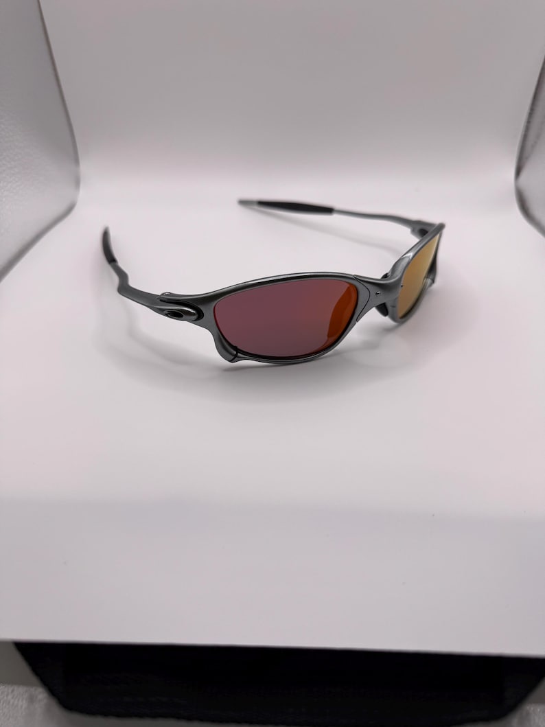 May include: A pair of sunglasses with a dark gray frame and tinted lenses. The lenses transition from a reddish-purple to a yellow-orange hue. The sunglasses are set against a white background.