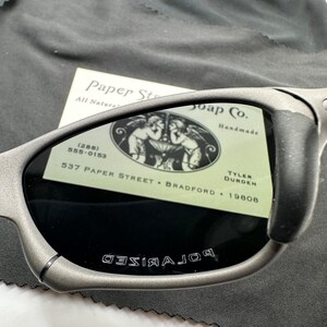 May include: Gray polarized sunglasses with the words "POLARIZED" etched on the frame. The sunglasses are resting on a gray microfiber cloth with a business card for Paper Street Soap Co. with the address 537 Paper Street, Bradford, 19808.