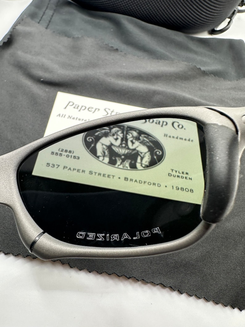 May include: Gray polarized sunglasses with the words "POLARIZED" etched on the frame. The sunglasses are resting on a gray microfiber cloth with a business card for Paper Street Soap Co. with the address 537 Paper Street, Bradford, 19808.