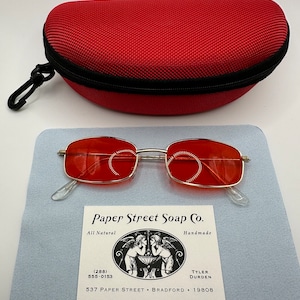May include: Red square-shaped sunglasses with a gold metal frame. The sunglasses are sitting on a light blue surface with a business card that reads "Paper Street Soap Co. All Natural Handmade (288) 555-0153 TYLER DURDEN 537 PAPER STREET BRADFORD 19808".