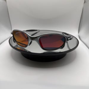 May include: A pair of sunglasses with a silver frame and gradient lenses, transitioning from orange to red. The sunglasses are displayed on a mirrored surface, reflecting the light and the frame's details. The overall design is sleek and modern.