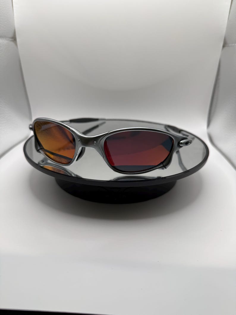 May include: A pair of sunglasses with a silver frame and gradient lenses, transitioning from orange to red. The sunglasses are displayed on a mirrored surface, reflecting the light and the frame's details. The overall design is sleek and modern.